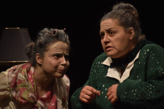 Nesta imagem estão em grande plano duas atrizes. Vê-se apenas do peito para cima. A da esquerda está ligeiramente curvada e usa uma camisa branca com um lenço floral. Tem rolos na parte de cima do cabelo grisalho e o resto está amarrado num puxo. O seu rosto tem rugas e encheu as bochechas de ar. A sua expressão é de gozo. A segunda atriz, no lado direito, tem o cabelo grisalho amarrado num puxo. Tem rugas no rosto. Veste uma blusa branca, um casaco verde escuro esburacado e um lenço no pescoço. Está com uma expressão assustada.