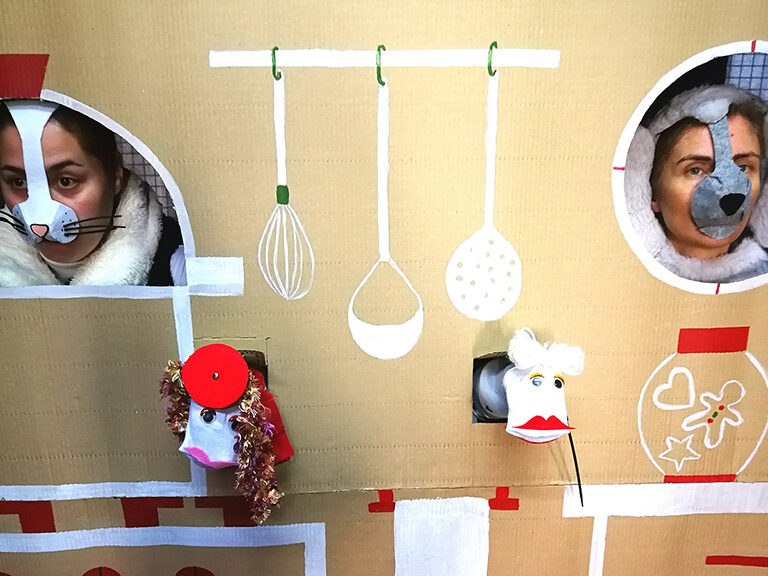 Esta imagem representa um pormenor de um cenário que é o interior de uma habitação. Duas atrizes espreitam através das janelas para o seu interior. A atriz do lado esquerdo tem uma máscara de gato e a atriz do lado direito tem uma máscara de cão. Entre elas espreitam também dois fantoches. O do lado esquerdo tem o rosto branco, nariz rosado, olhos escuros e pêlo encaracolado. Tem também uma boina vermelha. O do lado direito tem o rosto branco, lábio superior vermelho, um olho claro e outro escuro e cabelo curto branco.