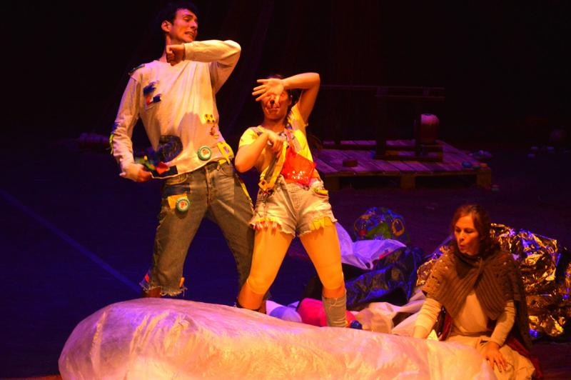 Nesta imagem estão um ator e duas atrizes. O ator tem vestido uma camisola branca e umas calças de ganga. Na sua roupa tem preso alguns objetos como um relógio redondo. Tem o braço esquerdo levantado e dobrado até ao peito. A atriz que está imediatamente à sua direita tem vestido uma t-shirt amarela, uns calções de ganga e umas leggings amarelas que vão até antes do joelho. Com o braço direito levantado e dobrado, bloqueia a cara. Ambos estão de pé. Mais à direita está uma senhora sentada num barco insuflável branco. A senhora veste um vestido branco comprido e um xaile cinzento sobre as costas e o peito. Atrás dos atores, está uma bola transparente insuflável e um monte prateado. No fundo do cenário, está uma palete de madeira pousada no chão.