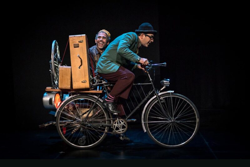 Imagem de cena do espetáculo "Car 12" onde aparece em primeiro plano um ator pedalando a sua bicicleta e em segundo plano o outro ator, com um ar risonho