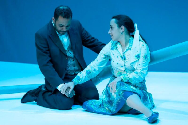 Imagem de cena do espetáculo onde estão os dois actores, um de joelhos e o outro sentado no chão, de mãos dadas
