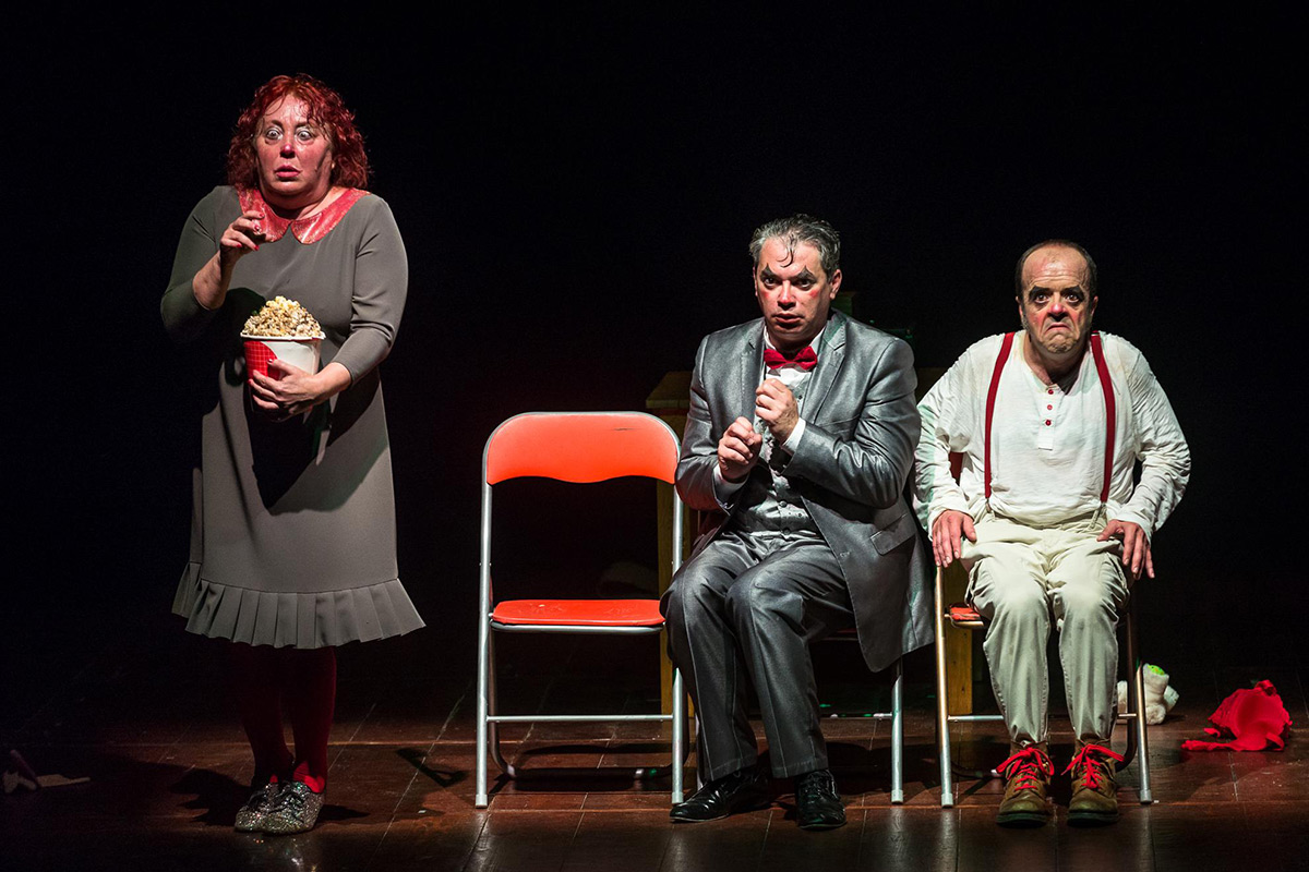 Plano geral de cena onde estão as três personagens. Os dois homens estão sentados à esquerda da personagem feminina que está em pé, havendo entre eles uma cadeira vermelha vazia.