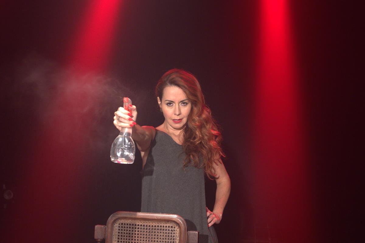 Sobre um fundo negro com dois feixes de luz vermelha, está, de pé, uma mulher vestida de castanho. O seu cabelo castanho e longo é, também, um pouco ondulado. Tem o braço direito esticado, para a frente e na mão, segura um pulverizador.