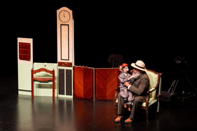 Palco de teatro com cen&aacute;rio de interior. &Agrave; direita, um homem sentado num cadeir&atilde;o segura um boneco vestido com fato cinzento e bon&eacute; vermelho. O homem usa chap&eacute;u claro e fato riscado. Ao centro, um biombo de madeira e uma estrutura com rel&oacute;gio no topo. &Agrave; esquerda, uma pequena mesa e um painel com um quadrado vermelho. Fundo preto.