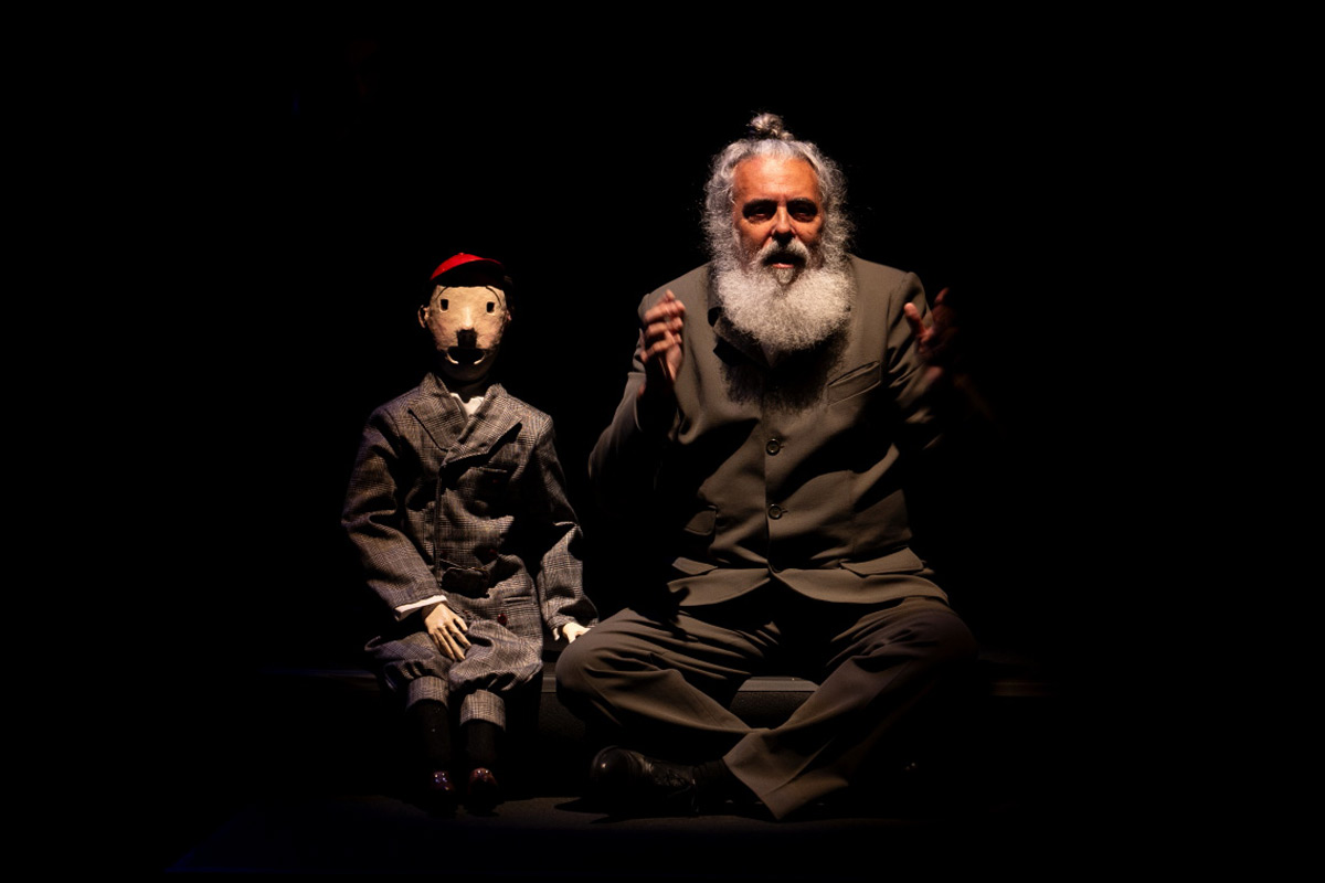 Um homem adulto, de meia-idade, com cabelos e barba grisalhos, está sentado em posição de meditação, num palco escuro. Ele veste um fato bege-acastanhado. Ao seu lado, um boneco, vestido de fato tom cinza-claro, está sentado. O boneco, com um chapéu vermelho, lembra uma criança. O homem apresenta-se no centro do enquadramento, enquanto o boneco está posicionado um pouco à esquerda. Ambos estão de frente para o observador. A composição é simples, com foco claro nos dois personagens, num fundo totalmente preto. A atmosfera é de um palco de teatro, ou peça de marionetes, com um tom sutilmente dramático.