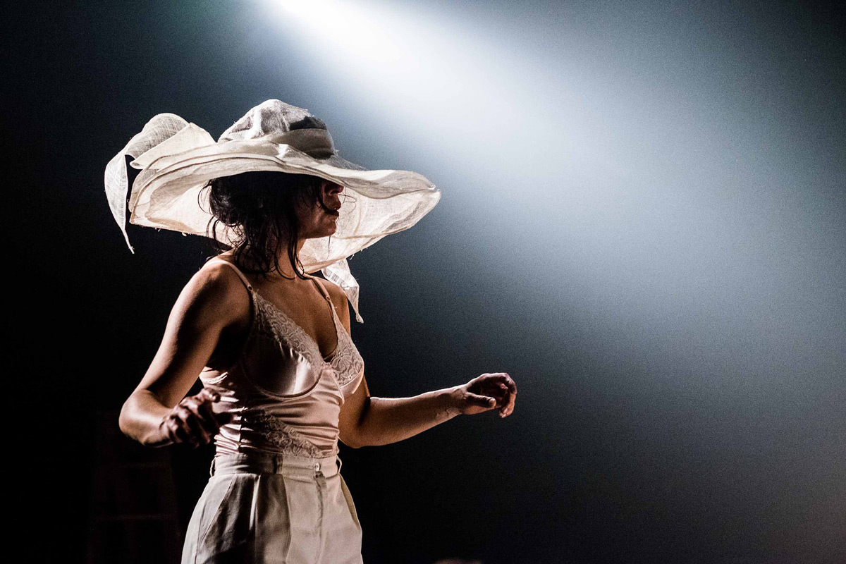 Uma mulher de pele morena e jovem, possivelmente entre 20 e 30 anos, está em um palco, em um ambiente escuro e com iluminação focada. Ela usa um chapéu de véu largo e branco, e um top de renda e calças de tecido leve, ambos em tom bege claro. Seu corpo está em um movimento dinâmico, os braços e mãos em uma pose que sugere expressão e dança. O cabelo é castanho escuro e está solto, caindo em ondas ao redor dos ombros. Sua expressão facial é subtilmente expressiva, com olhar voltado para o lado, para o espaço vazio ao fundo. A composição é na forma de um retrato de perfil, com foco na figura central que está ligeiramente posicionada à esquerda do quadro. O ambiente é um palco ou espaço cênico, com fundo escuro e um gradiente subtil de luz a partir de uma fonte de iluminação direcional, posicionada de cima para baixo, e de um lado para outro. A atmosfera é sugestiva de uma apresentação de dança ou performance.