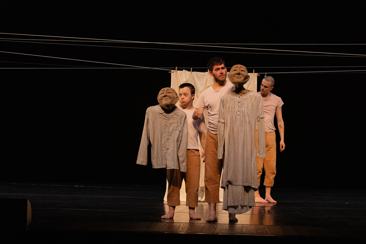 Cena de teatro com personagens vestidos com roupas de linho bege e castanho-claro, num palco escuro. Vários indivíduos, aparentemente adultos, com expressões faciais distintas, estão posicionados em formação, diante de uma pano branco pendurado num fio de aço. Os dois homens à esquerda seguram bonecos com máscaras faciais e roupas brancas, sem pernas. A iluminação é suave e focada nos personagens, destacando as texturas das peças de vestuário. A composição da imagem é simétrica e equilibrada, com os personagens organizados ao longo da área central do enquadramento. O fundo preto promove um foco direto nas figuras em cena. Os personagens parecem estar em um estado de contemplação ou movimento subtil. A perspectiva é de um espectador na frente do palco, com um foco claro nos figurinos e expressões dos personagens.