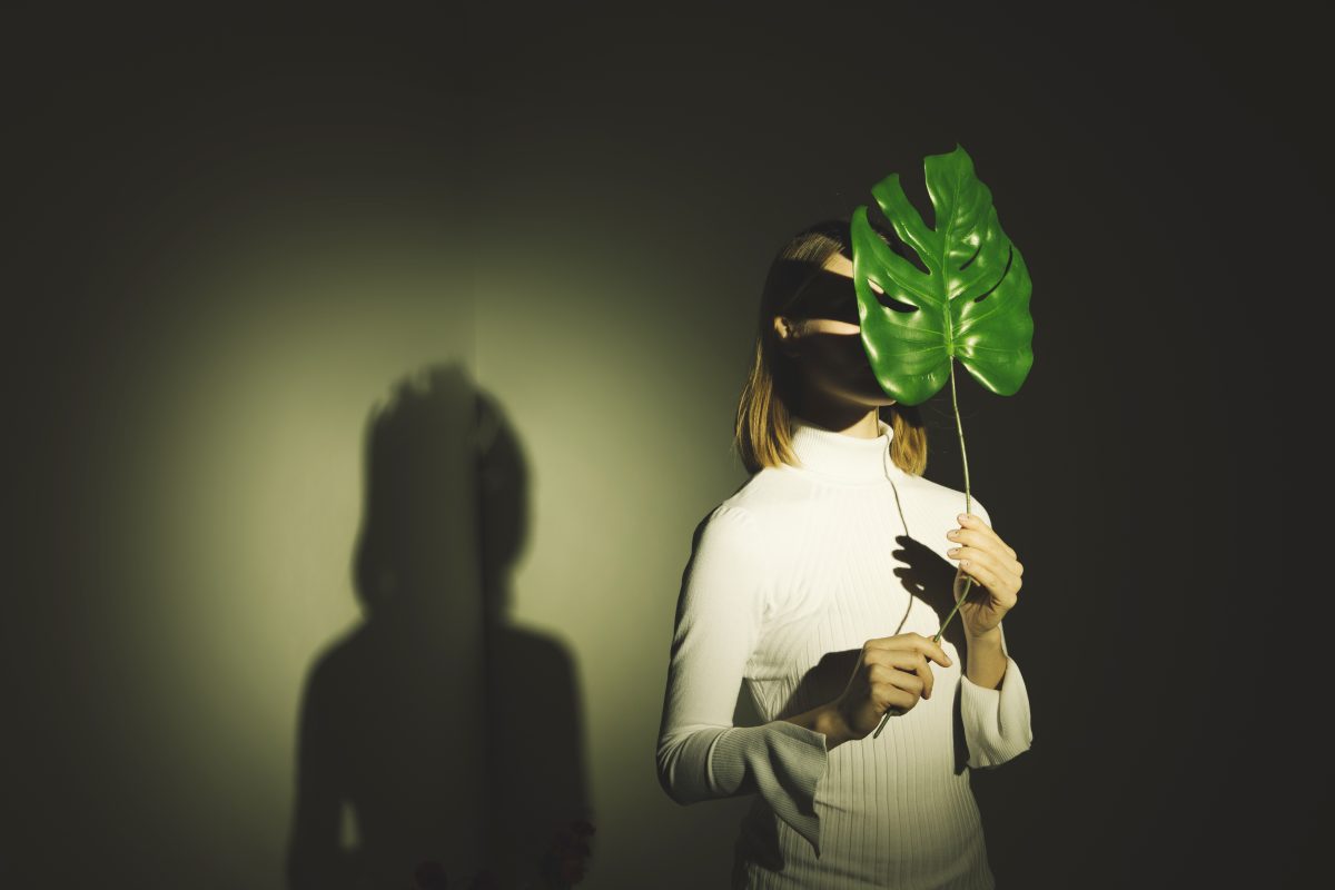 Uma mulher jovem, de pele clara e cabelo castanho curto, está à esquerda da imagem, em posição de três quartos de perfil. Segura uma folha verde-escura de planta tropical que cobre parcialmente o rosto. O olhar está voltado para o lado, não em direção ao observador.
Veste um suéter branco de gola alta. Não há outros acessórios visíveis. As mãos seguram a folha à frente do corpo.
O fundo é preto ou cinzento-escuro. A luz direta ilumina a mulher e a folha, projetando atrás dela uma sombra em tom amarelo-esverdeado.
A perspetiva é frontal, com o ponto de observação ligeiramente elevado em relação à mulher.